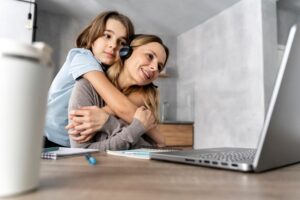 A mãe, uma mulher branca e loira, está sentada à mesa, usando fones de ouvido e com o notebook à sua frente, enquanto seu filho pequeno a abraça por trás. Ela sorri afetuosamente.