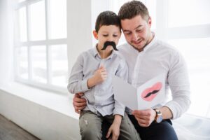 Pai e filho leem juntos um cartão de dia dos pais. O filho segura um bigode falso à frente do rosto.